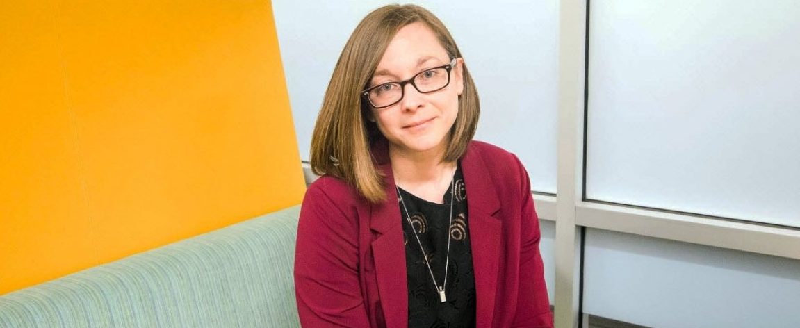 Trenia wears glasses and a black shirt with a magenta blazer. She is seated and looking at the camera, smiling slightly.